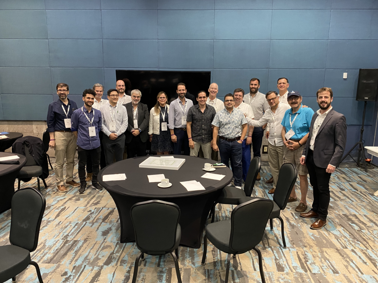A large group of people standing and smiling at the camera. They're in a blue room with many black round tables.