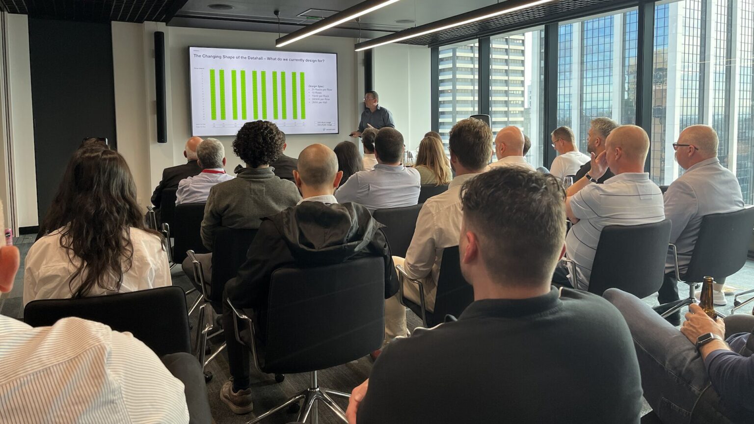 A large room of people face a man presenting on a projector. The screen shows a graph.